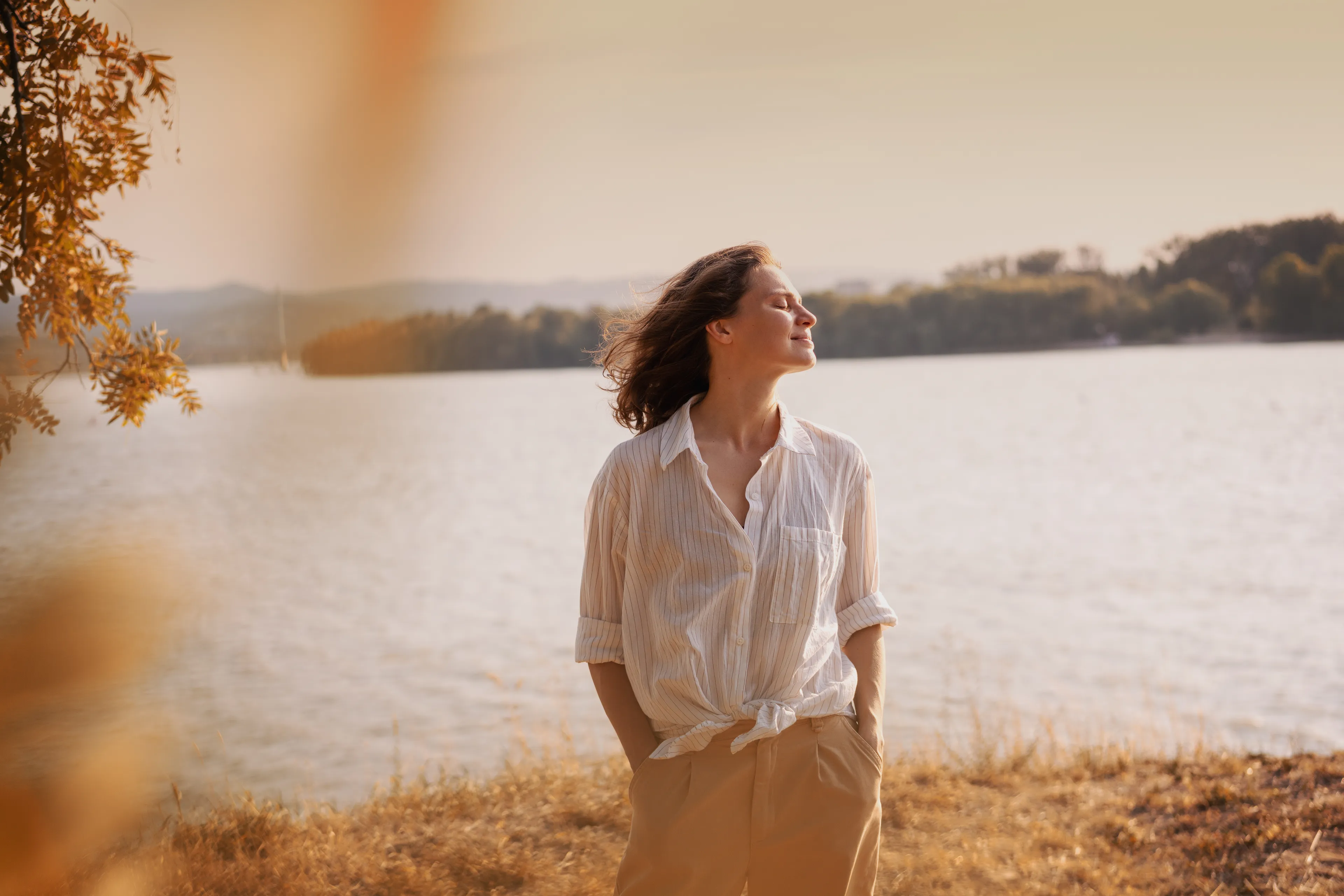 De vrouw op de foto staat voor een natuurgebied met water en lage heuvels. Ze kijkt naar rechts en heeft een uitgeruste, voldane uitdrukking op haar gezicht.
